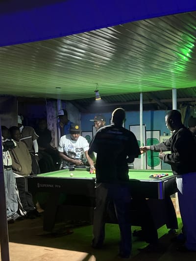 The image captures an indoor gathering where a group of men are playing pool. The setting appears to be a bar or a recreational space in Wumat, Nigeria. The room is illuminated by green and blue lighting, casting a vibrant and somewhat moody atmosphere. A pool table with green felt is the central focus, with billiard balls arranged in a rack and scattered on the table. Several individuals are actively engaged in the game, with cues in hand, while others observe or socialize. The overall impression is one of casual leisure and camaraderie.