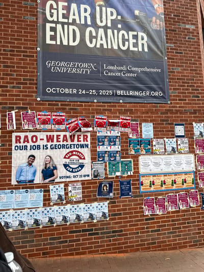 The image displays a densely covered reddish-brown brick wall on the campus of Georgetown University in Washington, D.C., United States, functioning as a public bulletin board. A large, dark blue banner at the top advertises the "GEAR UP. END CANCER." event for the Georgetown University Lombardi Comprehensive Cancer Center, scheduled for October 24-25, 2025, with a partial image of a cyclist. Below this banner, the wall is extensively covered with numerous paper posters and flyers of varying sizes, adhered haphazardly.
Prominently featured in the center is a large campaign poster for "RAO-WEAVER OUR JOB IS GEORGETOWN," displaying smiling photographs of a man and a woman, with "RAO-WEAVER OUR JOB IS GEORGETOWN FOR GUSA" and "VOTING: OCT 22 8PM" indicated. Surrounding this central poster are dozens of smaller flyers. Many repeat messages such as "MY BODY, MY CHOICE! Vote Ann Johnson" (pink/purple), "A BETTER WORLD... A BETTER GEORGETOWN" (red with QR codes), and various "Match-up" posters with candidate names and QR codes, likely related to student elections. Other visible posters announce events like "Rally for Gaza," "Gospel of John Bible Study," and provide information for the "Financial Accessibility and Equity Committee," or promote organizations like "The Philodemic Society." The ground at the bottom of the wall is paved, and a partial view of a dark object and a white bottle is visible in the lower left foreground.