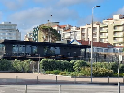 The image depicts a scene in Figueira da Foz, Portugal. In the foreground, a pedestrian crosswalk with visible white stripes and metal posts delineates the street. Beyond the crosswalk, a low hedge separates the street from a raised area with a building situated behind it. The building features a modern, dark-colored facade with large glass windows, possibly a restaurant or cafe. Behind the building, an array of multi-story buildings with various architectural styles and colors are visible, contributing to the urban landscape. The sky above is a clear blue with scattered clouds. A few lampposts are strategically positioned throughout the scene, adding to the overall urban setting. A volleyball net is erected at the front of the building, and a bike rack is visible to the left.