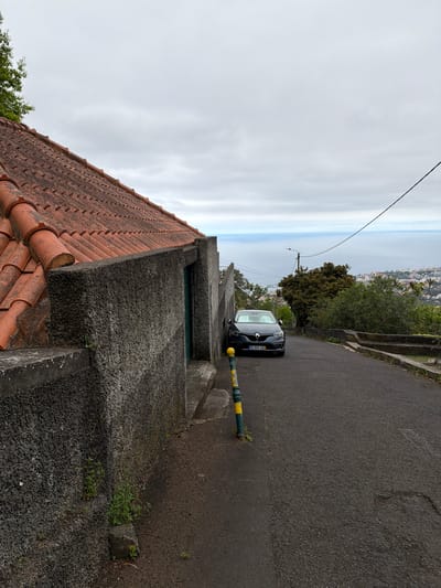 A black car is parked on the side of a narrow paved road in Funchal, Portugal. The road slopes downward into the distance, with a view of the city and ocean visible in the background. A low stone wall lines the side of the road, with a section of terracotta roof tiles visible in the foreground. A green and yellow bollard stands beside the car. The sky is overcast.