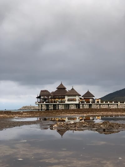 This horizontally oriented image features a grand building, likely a resort or hotel, situated by a calm body of water under an overcast sky. The multi-story building showcases distinctive tiered, peaked roofs, possibly resembling pagoda-style architecture, and appears to have several interconnected sections. Its facade is detailed with intricate latticework or decorative stone patterns, and numerous windows emit warm, golden light from within, contrasting with the muted natural light.

In the foreground, a rugged, muddy, or rocky shoreline or bank runs along the edge of the water. The tranquil water acts as a mirror, clearly reflecting the building's structure and its illuminated interior lights, creating a symmetrical visual effect.

Behind the building, a dark, undulating mountain range stretches across the horizon. The sky is a uniform expanse of grey, indicating an overcast day or perhaps dusk, contributing to a serene and somewhat subdued atmosphere. No people or visible text are present. The scene suggests a tranquil, potentially luxurious, setting, consistent with resort areas found in locations such as M'diq, Morocco.