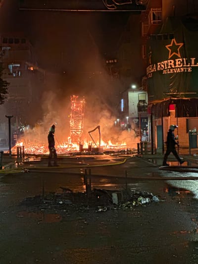 The image captures a scene at night in Alicante, Spain, during a significant fire. In the foreground, the wet asphalt reflects the surrounding light and flames. Debris, including plastic bottles and other refuse, is scattered on the ground. A yellow hose lies across the wet surface, suggesting ongoing firefighting efforts.
Further back, a large structure, appearing to be made of wood and possibly part of a "Falla" monument, is engulfed in intense flames, sending thick smoke into the dark sky. The fire illuminates the surrounding buildings.
Two individuals, presumably firefighters, are visible. One, in the mid-ground to the left of the burning structure, wears protective gear including a helmet and stands facing the fire. Another individual, to the right, also in protective gear, walks away from the camera, also appearing to be a firefighter.
On the right side of the image, a building is draped with a large green banner bearing a prominent star and text, partially obscured by smoke and the angle, but clearly visible is "ESTRELLA DE LEVANTE," indicating a prominent local brewery. Above this, balconies on a building show people observing the scene. The overall environment suggests a public area in Alicante experiencing a dramatic event.