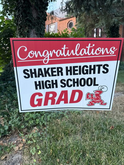 A celebratory yard sign for a graduate of Shaker Heights High School is displayed on a lawn. The sign features the text "Congratulations" in cursive white lettering on a red banner at the top. Below this, in bold black lettering, are the words "SHAKER HEIGHTS HIGH SCHOOL." The word "GRAD" is prominently displayed in large red capital letters at the bottom, accompanied by a red cartoon mascot with a chef's hat. The sign is supported by metal stakes and is situated in front of a brick house with a pitched roof and a visible circular window. Lush green trees and ivy are visible in the background, along with a patch of grass and some fallen leaves in the foreground. The scene appears to be outdoors in a residential setting.