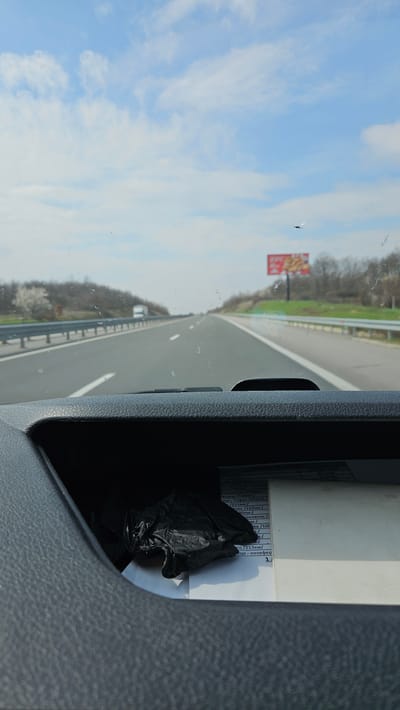 The image presents a forward-facing view from inside a vehicle onto a multi-lane highway in the vicinity of Malko Tranovo, Bulgaria. The asphalt road surface, marked with white lines, extends into the distance under a partly cloudy blue sky. Metal guardrails are visible on both sides of the highway, bordering treelines and embankments. A white truck is visible in the distance on the left lane. On the right, a large red billboard displaying an indistinguishable advertisement is situated beyond the guardrail. A small, dark, airborne object is visible in the upper portion of the sky through the windshield. The dark gray, textured dashboard of the vehicle occupies the foreground. An open compartment below the dashboard contains several white papers with Bulgarian text and a crumpled black object, consistent with a plastic bag or glove, resting on them. The windshield exhibits minor specks of dirt or dust.