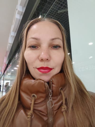A young woman with long, blonde hair pulled to the sides of her face is captured in a selfie. She is wearing a brown, puffy jacket with a zipped front and drawstrings on the hood. Her makeup includes bright red lipstick and mascara. She has fair skin with some freckles visible on her cheeks. Her eyes are a greenish-blue color.
The background appears to be an indoor setting, possibly a shopping mall or an indoor public space. To the left, there is a section of a building with architectural features, composed of white and grey rectangular elements. Beyond that, glass storefronts with blurred reflections of people can be seen. To the right, a large, light-colored wall is visible, with faint text in a Cyrillic script that appears to read "СВЯЗЬ" (meaning "connection" or "communication" in Russian). The lighting suggests it is daytime, with overhead lights visible in the background. The overall mood is casual and straightforward, a typical selfie taken in a public area. The woman's expression is neutral, and she is looking directly at the camera.
