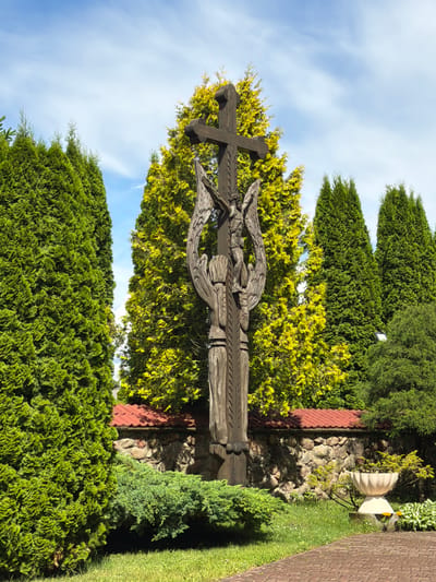 The image captures a tall, intricately carved wooden cross depicting Jesus Christ and two winged figures on either side. The cross stands outdoors, surrounded by lush greenery, including tall evergreen trees and smaller bushes. Behind the cross, a stone wall with a red-tiled roof is visible, suggesting it might be part of a religious or historical site. The sky above is blue with scattered white clouds. The scene appears serene and possibly contemplative. This sculpture is located in the vicinity of the Church of Saints Peter and Paul in Gerviaty, a village within the Gertsya village council of Belarus.