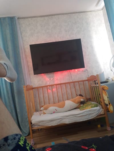A wide shot shows a bedroom in an apartment in Krasnovishersk, Russia, with a wooden baby crib in the foreground. The crib contains a white mattress and a long, brown and white stuffed animal resembling a dog. To the right of the crib is a gray dresser with a fan and other items on top. A large, flat-screen television is mounted on the wall above the crib, with red string lights running along the bottom edge of the TV. Blue curtains hang on the left side of the room. A person's legs, wearing dark blue and green patterned pants and a white sock, are visible in the bottom left corner of the frame. The floor is made of light-colored wood.