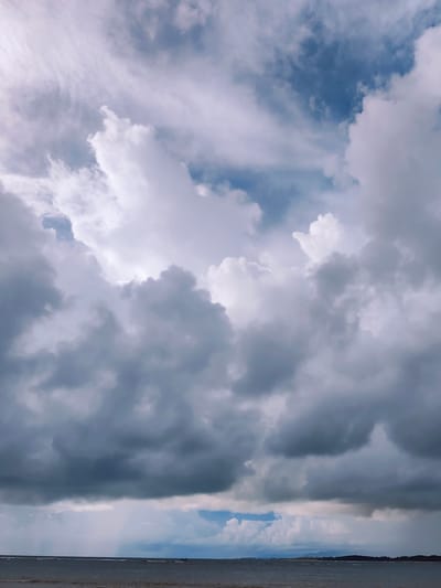 The image depicts a stormy sky over a body of water in Sakhu, Thailand. Dark, heavy clouds dominate the upper portion of the frame, with patches of lighter, wispy clouds and glimpses of blue sky peeking through. Below the dramatic clouds, the sea stretches out, appearing choppy with small waves. In the distance, a faint, dark line suggests land or a distant shoreline, possibly with some vegetation. The overall atmosphere is moody and intense, indicative of an approaching or ongoing storm. There are no visible people or distinct man-made objects in the scene, aside from a possible small boat far in the distance on the water.