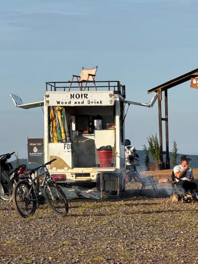 The image depicts an outdoor scene, possibly in Trường Thọ, Vietnam, featuring a food truck named "NOIR Wood and Drink." The food truck is white with several open windows and a metal railing on top. A folding chair is positioned on the railing. Inside the truck, colorful curtains are visible. Two bicycles are parked next to the truck, one black motorcycle is also present. A man in a white t-shirt and black baseball cap sits on a chair by a small campfire, holding a phone and looking down. He is positioned to the right of the food truck, with a wooden structure and greenery in the background. The ground is covered in gravel and sparse grass, and rolling hills are visible in the distance under a clear sky.