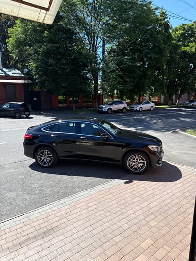 An eye-level, full shot captures a black Mercedes-Benz GLC Coupe parked on the right side of a paved city street. The car is angled towards the camera, with its front-right wheel prominently displayed. The vehicle is a modern coupe SUV with a sleek, black exterior and chrome accents.

The street is a mix of asphalt and a brick-patterned sidewalk. The sidewalk runs along the right edge of the frame and is made of light-colored rectangular pavers. The asphalt road appears to be a two-lane street with clear white lane markings.

Behind the Mercedes, other vehicles are visible on the street. Further down the road, a silver SUV and a white sedan are parked. To the left of the Mercedes, a smaller black SUV is parked near a brick building.

The background is dominated by lush green trees, indicating a well-maintained urban or suburban environment. The sky is a clear, bright blue. The scene is bathed in bright sunlight, casting shadows from the trees and the car onto the pavement. The overall impression is of a bright, sunny day in a city.