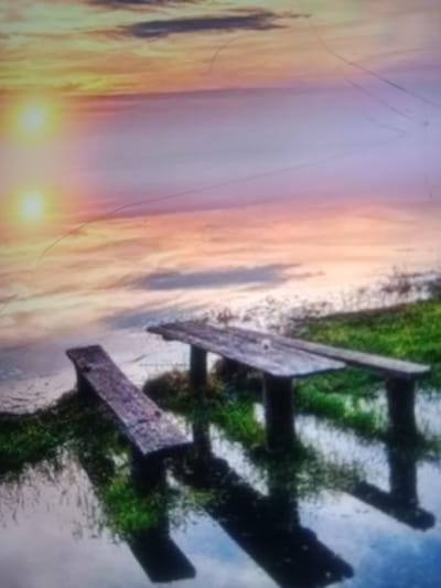 The image depicts a serene waterscape at dusk with two wooden benches situated on a grassy bank. The sky displays a gradient of warm colors, transitioning from yellow and orange near the horizon to pink and purple at the top, suggesting a sunset. Two circular, bright orbs, likely reflections of the sun or lights, are visible in the sky on the left side. The water in the foreground is calm and reflective, mirroring the sky and the benches. The benches appear weathered and aged, with one positioned at an angle closer to the viewer and another further back, appearing as a table-like structure with two upright supports. The surrounding environment is characterized by lush green grass. There are no people or animals present in the scene. The overall atmosphere is one of tranquility and natural beauty, potentially at a lakeside or riverbank. The image is grainy and exhibits some scratches or artifacts, possibly due to its capture or display medium. No specific landmarks or architectural elements are identifiable to pinpoint a location within Kielce, Poland.