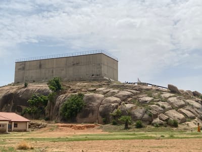 The image shows a large, rectangular, concrete reservoir structure atop a rocky hill in Kufena, Nigeria. The reservoir has a metal railing around its top perimeter. A pipeline runs along the rocky ground behind the reservoir. The hillside is covered with large, rounded rocks and sparse vegetation, including trees and palm-like plants. At the base of the hill is a small building with a red roof. The foreground consists of dry, sandy ground interspersed with patches of green grass. In the lower right corner, two figures appear to be walking, one wearing an orange robe. The sky is partly cloudy.