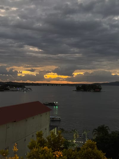 A river scene under a dramatic sky filled with dark, swirling clouds contrasted by a band of golden sunlight breaking through. The water reflects the somber mood, punctuated by the lights of distant structures along the shoreline. A building with a red roof and a bush occupies the lower right corner. Several boats or ferries are visible on the river. A few palm trees with lights are on the right side of the photo.