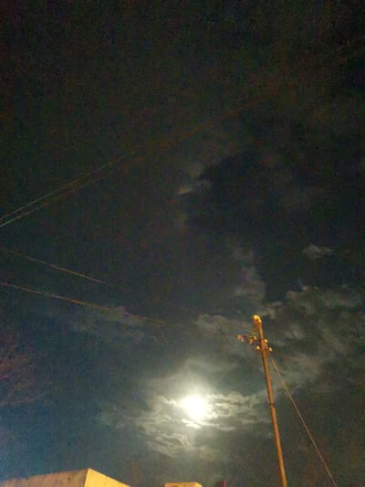 A nighttime scene captured from what appears to be a lower elevation, looking upwards towards a dark, cloudy sky. The moon, partially obscured by clouds, is a bright focal point in the lower center of the image. A wooden utility pole, topped with insulators, stands prominently on the right side of the frame, with wires extending from it and across the sky. A portion of a building, with a light-colored facade, is visible in the lower left corner. The image quality is grainy, suggesting it was taken in low light conditions. This scene was captured in the city of Obligado, Paraguay.