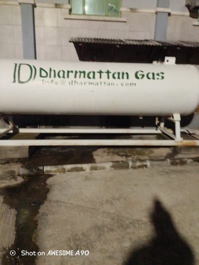 The image displays a large white gas cylinder labeled "ID Dharmattan Gas" with the email address "info@dharmattan.com" printed below it. The cylinder is supported by a white metal frame situated on a concrete surface. The background shows a building with light-colored tiled walls and a dark-colored corrugated roof structure above the cylinder. The lighting suggests it is nighttime, with a shadow of a person visible in the lower right quadrant of the image. The "Shot on AWESOME A90" watermark in the bottom left corner indicates the device used to capture the photograph. The overall scene is outdoor, likely a commercial or industrial area, and could be located in Ibadan, Nigeria, given the context of the prompt.