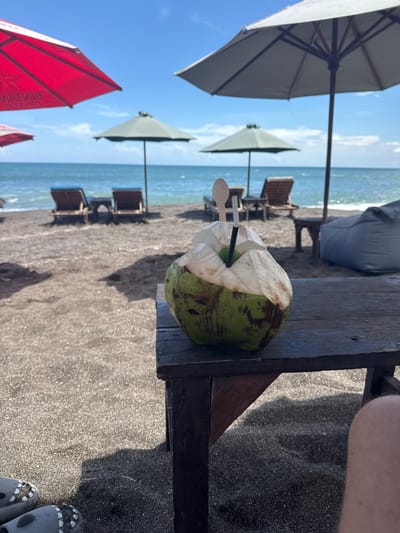 This image was taken on a beach in Canggu, Indonesia. The scene shows a young coconut with a straw and spoon in it sitting on a wooden table on the beach. There are several beach umbrellas and lounge chairs behind the table, with the ocean visible in the background. A person is standing next to the table with their feet and shoes in the lower right corner. The beach has gray sand. The weather appears to be sunny.