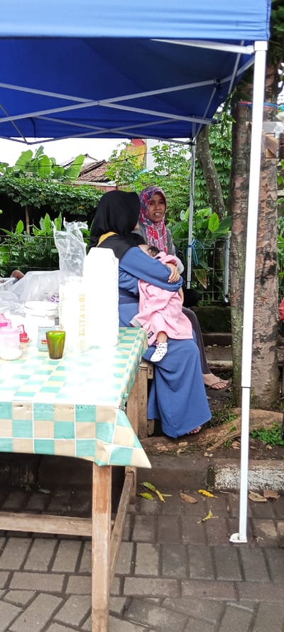 The scene takes place in Banjaran, Indonesia, specifically at the coordinates -7.046 latitude and 107.581 longitude. A woman is sitting at a table with a checkered tablecloth, holding a young child in her arms. The child is wrapped in a pink blanket and wearing pink pants. The woman is wearing a blue head covering and a blue dress. A second woman wearing a patterned head covering and a pink and red shirt is seated next to her, looking towards the camera. The women are seated under a blue canopy with a white frame. The table is covered in a checkered tablecloth and includes a plastic bag with the label "ZETTA 10 OZ", a white bowl, a glass container, a small pink bottle, and a green glass. The ground is a brick walkway and the surrounding area is made up of brick walls and a few plants and trees. A white metal pole is part of the canopy's structure. The scene has an informal and relaxed feel. The women are interacting with each other while the child remains calm in the woman's arms. The location is most likely an outdoor market or food stall.