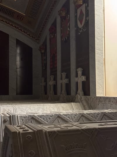 The image depicts the Pantheon of Kings in the Monastery of San Lorenzo de El Escorial, Spain. The scene shows a dimly lit interior with marble walls and sarcophagi. In the foreground, ornate sarcophagi with intricate carvings are visible. Behind them, several marble crosses stand on pedestals. The background features dark panels adorned with coats of arms and crowns, likely commemorating royalty. The overall atmosphere is solemn and historical, reflecting the grandeur of the royal burial site.
