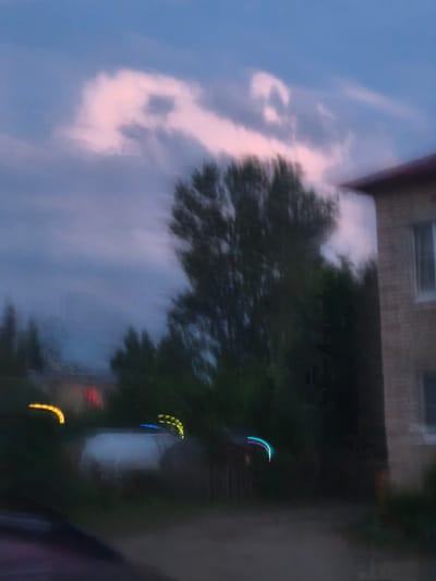 The image depicts an abstract, impressionistic scene with motion blur, suggesting a late evening or twilight setting. A large tree dominates the mid-ground, its form blurred against a dusky sky streaked with pink and blue clouds. To the right, the corner of a brick building with a reddish-brown roof is partially visible, featuring a window. In the foreground, several streaks of colored light, appearing as yellow, blue, and red, suggest movement, possibly from vehicles or streetlights. The lower portion of the image shows a darker, unidentifiable ground surface. The location is identified as Kady, Russia, though specific landmarks are obscured by the motion blur.