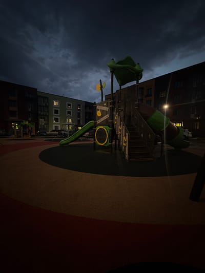 The image shows a children's playground located in the city of Фролы, Russia, at dusk or night. The playground features a green tube slide, a set of wooden stairs leading up to it, and a climbing net structure. The ground of the playground is covered with dark, textured material, with a circular section in the foreground that is a mix of red and yellow. In the background, apartment buildings with lit windows are visible. The sky is dark with heavy clouds. There are no people or animals present in the image.