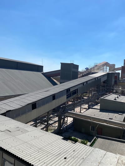 The image shows a tall industrial building, possibly a factory or a power plant, against a clear blue sky. The building appears to be under construction or renovation, with scaffolding visible around some sections. There are several large, corrugated metal structures that form part of the building's exterior. Some pipes and other industrial elements are also visible.

In the middle distance, a few people can be seen working on the scaffolding, wearing safety helmets. The scene suggests an active industrial environment. The sun is shining brightly, indicating it's daytime, likely late morning or early afternoon.

The overall impression is one of a large-scale industrial complex, possibly in a rural or semi-industrial area, given the clear sky and the absence of other prominent structures. The weather appears to be good, with no signs of rain or clouds. There is no visible text in the image. The location context provided indicates Poland.