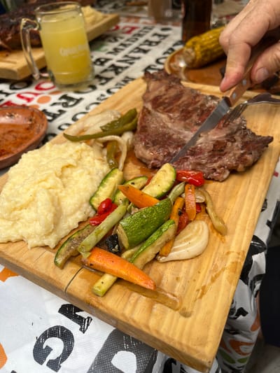 A wooden cutting board holds a grilled steak, sauteed vegetables (zucchini, carrots, peppers, and onions), and mashed potatoes. A person is using a fork and knife to cut the steak. A glass of yellow liquid sits beside the cutting board. The setting appears to be a table in a restaurant or home. The environment contains tablecloths with bold prints. It is difficult to determine the location.