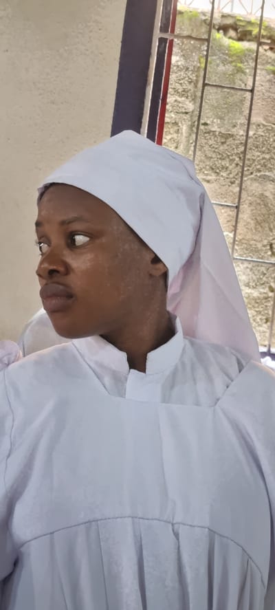 The image, taken in Ijegun, Nigeria, shows a close-up, rotated view of a young Black person wearing a white religious garment and head covering. The subject's face is partially visible, with their head tilted down and to the side, their gaze directed downwards and to the left. Their skin shows some patches of lighter pigmentation, particularly on the forehead and cheeks. The white garment appears to be a robe or cassock with a cowl or hood, made of a light fabric with visible pleating and seams.

In the background, a wall made of rough-hewn stone blocks is visible, with some patches of green moss. Horizontal bars, possibly part of a window or grate, run across the top of the stone wall. Below the stone wall, a ledge or window sill is visible, with a dark blue band and a thinner red band along its edge. The foreground wall is a light, textured color. The lighting suggests it is daytime, with natural light illuminating the scene. The overall impression is one of quiet contemplation or religious observance. There is no visible text in the image.
