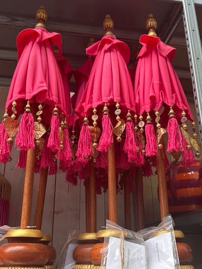 In a retail setting, possibly a market stall or shop, in Tuk, Netherlands, several vibrantly colored pink ceremonial umbrellas are displayed. The umbrellas are tightly furled and adorned with intricate gold-colored charms and pink tassels hanging from their edges. Each umbrella features a wooden pole topped with a decorative golden finial. The bases of the umbrellas are made of dark wood with a golden rim. In the background, a metal shelf unit is visible, and parts of other items, including a striped orange and purple container, are present. The scene is well-lit, highlighting the rich colors and textures of the merchandise.
