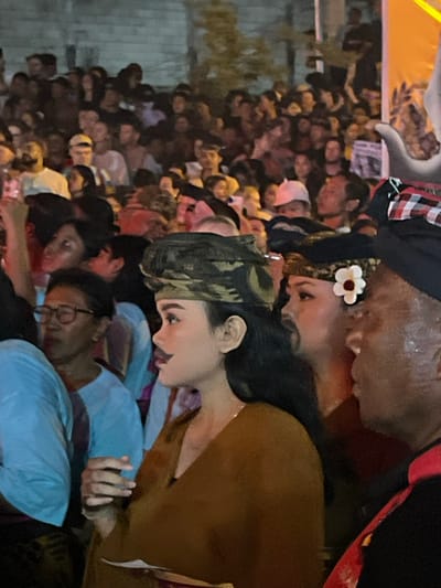 The image depicts a crowded outdoor nighttime scene in Munggu, Indonesia. In the foreground, a woman with dark hair wears a camouflage-patterned headwrap, a brown garment, and has a painted mustache and red lipstick; she holds a small white object in her right hand and looks toward the right. Behind her and slightly to the right, another woman is partially visible, wearing a dark headwrap with a white flower embellishment. To the far right, a man with dark skin, wearing a traditional red and white checkered Balinese udeng and a black and red garment, is partially visible, facing left. To the left, a woman wearing glasses and a light blue top observes the scene. The background is filled with a dense, out-of-focus crowd of people under artificial lighting, suggesting a public event or performance.