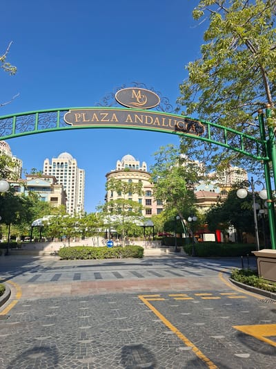 The image depicts the entrance to Plaza Andalusia in the Qanat Quartier area of Doha, Qatar. An ornate green archway with decorative swirls frames the view, bearing a dark sign with "PLAZA ANDALUSIA" inscribed in gold lettering and a circular emblem above it featuring the intertwined letters "M" and "U". Beyond the arch, a paved walkway with a cobblestone pattern leads towards several multi-story buildings characterized by Mediterranean architectural styles, including domed roofs and arched windows. Lush green trees and manicured bushes are strategically placed throughout the scene, with some bearing white flowers. Pedestrian crossings are marked on the paving. The sky is a clear, vibrant blue, indicating a sunny day. There are no people visible in the image. The overall scene suggests a well-maintained and picturesque public space designed for pedestrian enjoyment.