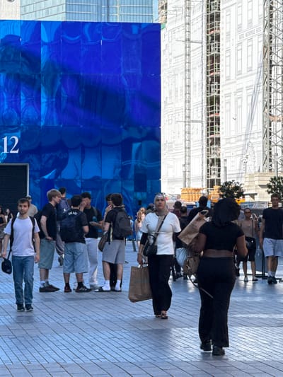 The image depicts a group of people standing on a patterned paved surface, likely a pedestrian walkway or plaza, in Brussels, Belgium. The setting appears to be urban, with a large blue tiled structure on the left and a building facade with intricate architectural drawings on the right, suggesting a construction site or an art installation. The time of day appears to be daytime, with ample sunlight casting shadows, indicating clear weather.
The people are casually dressed, and their poses suggest they are either waiting, observing, or conversing. Some individuals are holding bags, and one person is wearing a backpack. There is no clear indication of a specific event, but the gathering of people in an urban public space is evident.
Notable details include the architectural drawings on the building, which seem to depict a hotel with the text "HOTEL METROPOLE HOTEL" visible. The blue tiled structure has a large number "12" printed on it. The overall mood seems neutral, with no strong emotional expressions visible. The orientation of the image is such that the people are facing the blue wall and the building facade.
