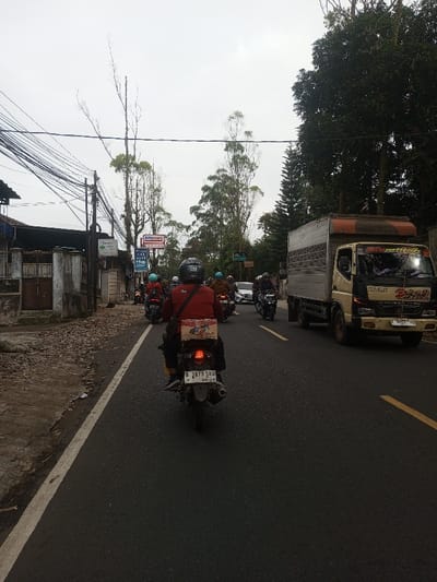 The image depicts a busy road scene under an overcast sky in Sat Thay (East), Myanmar. In the foreground, a dark asphalt road features a solid white line on the left shoulder and a broken yellow line indicating traffic lanes. A person wearing a black helmet and a red jacket is riding a black motorcycle, viewed from behind. A cardboard box, labeled "Indomie Goreng," is secured to the rear of the motorcycle. The illuminated taillight is visible, as is the license plate "B 3879 SKU" with "09-28" below it.
Ahead of this motorcycle, several other individuals are riding motorcycles in the same direction, wearing various colored helmets (black, blue, brown) and jackets. To the far left, a dirt and gravel shoulder transitions to a sidewalk bordering a residential-style structure with a brown gate and stone pillars. Utility poles laden with numerous black cables line the left side of the road. Visible signs include "Alfamart" and "Sinar Tani," along with other partially obscured commercial signage.
To the right, a beige and black box truck with an orange-brown top and visible decals is proceeding in the opposite direction, its headlights illuminated. Further down the road on the right, a white car is partially visible behind the truck. The right side of the road is bordered by dense, tall green trees, and the sky above is a uniform light gray.