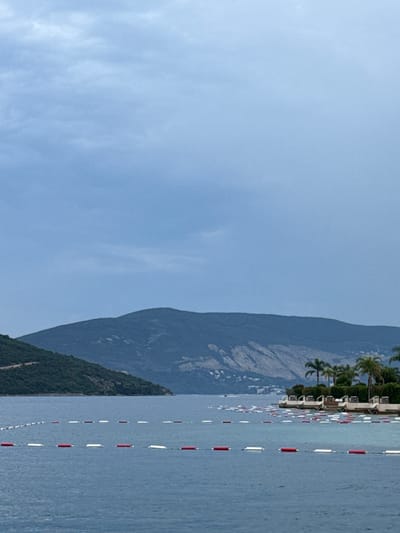 This media file depicts a serene coastal scene with a prominent mountain backdrop. The water, a tranquil blue, is marked by buoys with alternating red and white floats, indicating a designated swimming area or a marker for boats. Palm trees and lush greenery adorn the shoreline, suggesting a tropical or Mediterranean climate. Several white structures, possibly buildings or part of a resort, are visible along the coast, interspersed with more palm trees. The sky is a clear, pale blue with a few wispy clouds, indicative of fair weather and likely daytime. The overall atmosphere is peaceful and picturesque, evoking a sense of relaxation and vacation. The mountainous terrain hints at a dramatic, natural landscape typical of coastal regions. No people are visible, nor is there any text present in the image. The lighting suggests it is likely late morning or early afternoon.