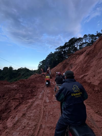 In Nao Sẻ, Vietnam, a dirt road carved through a recently excavated hillside is occupied by several motorcycles and construction equipment. The ground is a deep reddish-brown mud, heavily rutted by the tracks of heavy machinery, indicating ongoing construction or landslide remediation. The sky above is a gradient of blues and grays, with scattered clouds suggesting it is either early morning or late evening.
In the foreground, a person is riding a motorcycle, facing away from the camera. They are wearing a dark blue jacket with a yellow logo that reads "Vemelin Corporation." Behind this rider, two other motorcycles are visible, carrying passengers. The second motorcycle has a rider wearing a red helmet and a passenger in a yellow jacket with a red backpack. The third motorcycle has a rider wearing a white cap.
Further down the muddy track, two large excavators are actively working. One is a red Hitachi excavator, and another appears to be a similar construction machine. Trees line the top of the hill to the left and right of the road. The overall scene depicts a challenging and active construction site in a rural Vietnamese setting.