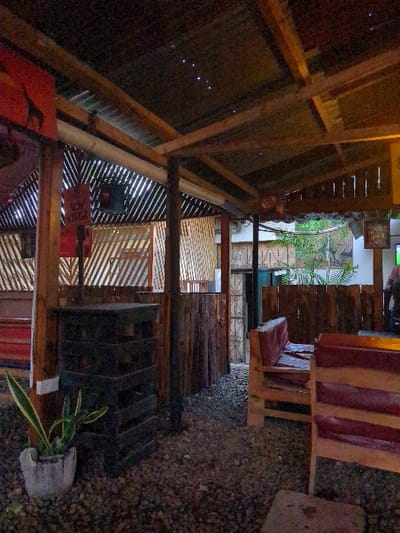The scene is a partially enclosed, wooden structure with a corrugated metal roof. It is located at coordinates -6.835, 37.653 in Morogoro, Tanzania. The structure is situated within a gravel yard, which is partially shaded by the structure itself. The yard is surrounded by a wooden fence and a metal gate. The wooden structure contains a table and several chairs. There are two people sitting at the table, one in a chair and one on the floor. There are two plants in the yard. The structure is lit by a string of lights. There is a sign hanging on the wall that says "VIP KING2." There is a door to the right of the table, through which you can see a metal gate. The yard is surrounded by a wooden fence. The scene is peaceful and relaxing.