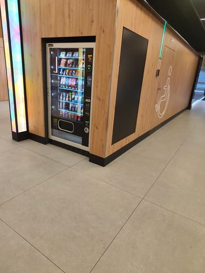 The image shows a vending machine and a section of a wall with a door and a wheelchair symbol. The vending machine is filled with various snacks and drinks. To the left of the vending machine, there are vertical illuminated panels with abstract colorful patterns. The wall is made of wood-look material and has a black rectangular panel on it, likely a door or an access panel. Above the wall, there is a green light strip. A white stylized wheelchair symbol is visible on the wooden wall, indicating accessibility. Below the wheelchair symbol, there is a small button and text that appears to say "WOLNE ZAJETE," which translates to "FREE OCCUPIED," suggesting this area might be related to accessible facilities or restrooms. The floor is tiled with large, light-colored square tiles. The overall scene appears to be in an indoor public space, possibly a shopping mall, train station, or a similar facility in the city of Łódź, Poland, given the Polish text. There are no people visible in the image.