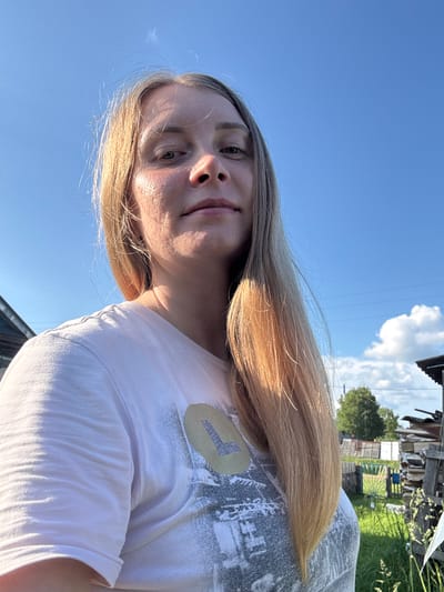 An outdoor, eye-level, close-up, selfie-style shot shows a fair-skinned woman with long, blonde hair and light freckles across her nose and cheeks. She is wearing a light pink t-shirt with a distressed, light gray graphic and a circular emblem with a yellow "L" on the left side of her chest. She is outdoors in front of a bright blue sky with a few wispy white clouds. In the background, several structures are visible, including a wooden building to the left and stacks of lumber and firewood to the right. Green grass and trees are also present. The overall setting appears to be rural, possibly a dacha or countryside home, consistent with a location in or near Kurgan, Russia.