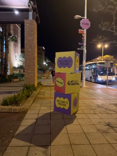 Captured at night in Punta Umbría, Spain, the image focuses on a stack of three large, colorful cubes placed on a paved pedestrian walkway. The cubes are decorated with text and symbols, including "WELCOME TO PARADISE," a smiley face, a starburst, and the text "LA *wtf* PUNTA." A bus is partially visible in the background, illuminated by its headlights and streetlights. To the left, a portion of a brick building and palm trees are discernible. The walkway is lined with a low brick and green vegetation border. A light post with a pink sign is partially visible. The lighting suggests an evening setting, with shadows cast by the cubes onto the walkway.