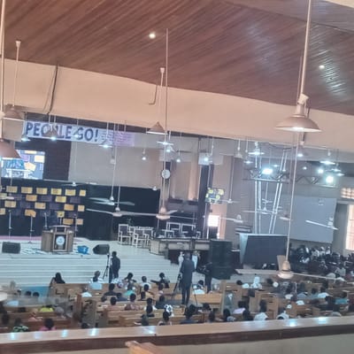 The image depicts the interior of a church in Jama A, Nigeria. The church is filled with people seated in wooden pews. At the front of the church is a raised stage with a pulpit and a backdrop of geometric shapes. A banner reading "PEOPLE GO!" is hung above the stage. Several ceiling fans and pendant lights hang from the ceiling. People are watching the service, and some are filming. Two cameramen with tripods are present on the steps in front of the stage. A clock is visible on the wall behind the stage. Several monitors are positioned around the stage.