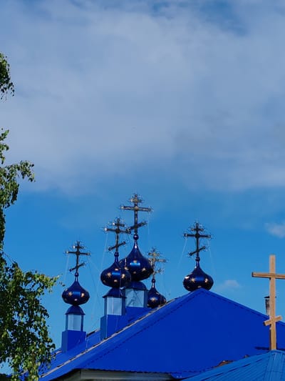 The image shows the blue domes and crosses of a Russian Orthodox church, likely located in the city of Chukhloma, Russia. The domes are dark blue and adorned with star-shaped gold accents. Each dome is topped with a cross, some of which are ornate and others are simpler. A large wooden cross is visible on the right side of the frame, placed on a smaller roof structure. The roof of the church is also a vibrant blue. On the left side of the image, the branches of a leafy green tree extend into the frame. The sky behind the church is a bright blue with scattered white clouds. The overall scene suggests a sunny day and a religious structure with distinctive architectural features common to Russian Orthodox churches.