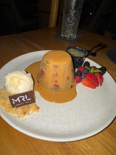 A meticulously plated dessert is presented on a white speckled ceramic plate, resting on a wooden table. The central subject is a cylindrical, possibly cake-like item, generously covered in a smooth, light caramel-colored sauce. To its left, two scoops of vanilla ice cream are positioned on a bed of golden-brown crumbles. Embedded within these crumbles is a small, rectangular dark chocolate plaque displaying "MRL" in white stylized letters, with "GRUPO MRL" printed beneath. To the right of the central dessert, an assortment of fresh berries, including whole raspberries, blueberries, and sliced strawberries, is neatly arranged and garnished with a sprig of mint. Behind the berries, a small, black speckled ceramic cup holds a pale, possibly viscous liquid. In the background, a tall, clear glass with a decorative pattern, containing liquid, sits on a dark coaster. A dark metallic object, resembling tongs or a utensil, is partially visible behind the black cup. The scene suggests an indoor restaurant setting, characterized by dim lighting and wooden furnishings, belonging to Grupo MRL in León, Guanajuato, Mexico.