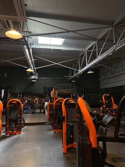 This image depicts the interior of a Basic-Fit gym in Alès, France. The scene is dominated by rows of orange and black weight machines, characteristic of the Basic-Fit brand. The gym features high ceilings with exposed metal beams and industrial lighting fixtures. Natural light filters in through skylights. Several individuals are visible engaged in various activities: a young man is seated on a machine, appearing to exercise; a woman is standing near a weight rack, possibly resting or waiting. Another person is seen walking in the background. Mirrors line one wall, reflecting more of the gym equipment. The flooring is dark and appears to be a durable, non-slip material. The overall impression is that of a functional, modern fitness facility.
