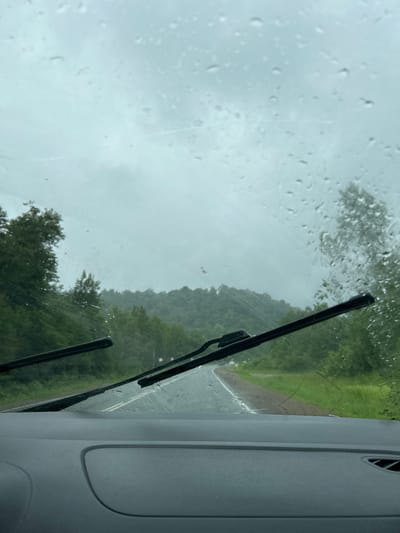 The image shows a view from inside a vehicle looking out through a rain-streaked windshield. Two windshield wipers are visible, one on the left and one on the right, partially clearing the view. The road ahead is wet and appears to curve to the right, surrounded by lush green trees and rolling hills that are shrouded in a misty, overcast sky. The overall impression is one of a rainy day drive through a rural, forested landscape. There is no indication of the city of Кулмас, Russia in the image.