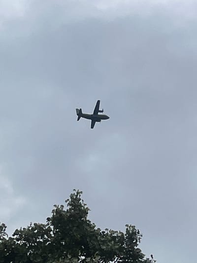 The image shows a twin-engine propeller airplane flying in a cloudy sky. The airplane is silhouetted against the overcast sky, with its wings and engines clearly visible. The bottom of the airplane has two smaller wings or stabilizers. To the right of the airplane, a portion of a green leafy tree is visible, suggesting that the photo was taken from the ground. The overall mood of the image is somewhat somber due to the cloudy weather, with the airplane being the central object of interest. The time of day is not explicitly indicated, but the overcast sky suggests it could be daytime. There is no visible text in the image. The location context of Cheshnegirovo, Bulgaria, places this scene in a rural or semi-rural setting. The activity is simply the airplane in flight.
