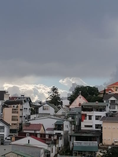 This image depicts a dense urban landscape with a variety of buildings, likely residential, nestled among rolling hills. The architecture suggests a European colonial influence, with multi-story structures featuring pitched roofs, balconies, and various window styles. The scene is dominated by a dramatic sky filled with dark, heavy clouds, hinting at an impending storm. In the foreground, a cluster of buildings exhibits a mix of architectural styles and states of repair. Some are painted in muted colors, while others feature more vibrant hues like orange and red on their roofs. A prominent pink building with a cross at its apex, possibly a church or religious institution, stands out amongst the other structures. The surrounding vegetation is limited, with a few trees visible, one of which is a tall, slender pine. The overall impression is one of a densely populated hillside town, possibly experiencing inclement weather. The presence of structures and architectural elements suggests a location within Lang Biang - Da Lat, Vietnam, a region known for its French colonial architecture and hilly terrain.