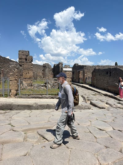 The photograph shows a man standing on a cobblestone street in Pompeii, Italy, adjacent to ancient Roman ruins. The man is wearing a blue cap, a light-colored button-down shirt, gray pants, and sandals. He has a camera hanging around his neck and a backpack.

Behind him, there are remnants of stone walls, suggesting the outline of former buildings. The ruins are weathered, revealing their age and the destructive impact of Vesuvius. A metal fence is in place, presumably to protect the site.

The sky is visible above the ruins, showcasing a blue sky with scattered white clouds. Two figures stand in the background. The scene suggests a typical day of tourism and exploration at this historical site.