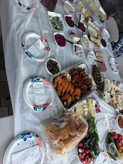 A spread of food is laid out on a white plastic tablecloth, likely for an outdoor gathering. The food includes several dishes of chopped vegetables, dips, cheeses, olives, pickles, sliced meats, bread, and a large platter of what appears to be grilled salmon and shrimp. There are also plates, cups, and cutlery set out. In the background, partially visible, is a person wearing a camouflage pattern shirt. The environment suggests a residential backyard or patio area. This scene is located in Covina, California, United States of America.
