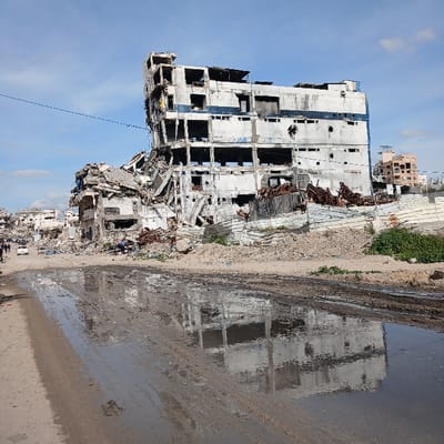 The image depicts a scene of extensive structural destruction in Jabalia, Palestinian Territory, under a partially cloudy blue sky. In the center, a severely damaged multi-story concrete building stands, its facade largely blown out, revealing internal floors and exposed rebar. The building, originally light-colored with blue accents, is heavily soiled and fragmented, with large sections of its walls and roof missing or collapsed. To the left, another collapsed structure is visible, surrounded by immense piles of concrete rubble, twisted metal, and general debris extending into the background, where more damaged buildings are discernible. In the far right background, a less damaged multi-story building with an orange-brown facade is partially visible. The foreground features an unpaved, muddy road with large puddles reflecting the damaged structures and the sky. On the left side of the road, in the midground, several dark figures, likely individuals, are observed near a white sedan. Sparse green vegetation grows amidst the rubble on the right. A thin, dark line, possibly a utility cable, stretches across the upper portion of the frame.