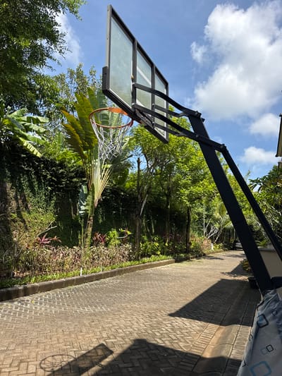 The image depicts a residential scene, likely in Jimbaran, Indonesia given the vegetation. A basketball hoop and backboard dominate the left side of the image, positioned near a paved driveway that extends into a lush garden area. The garden contains a variety of tropical plants, including palm trees and other greenery characteristic of the region. Above, a blue sky with scattered clouds is visible. The setting suggests a private residence with outdoor recreational space and landscaping typical of a tropical environment.