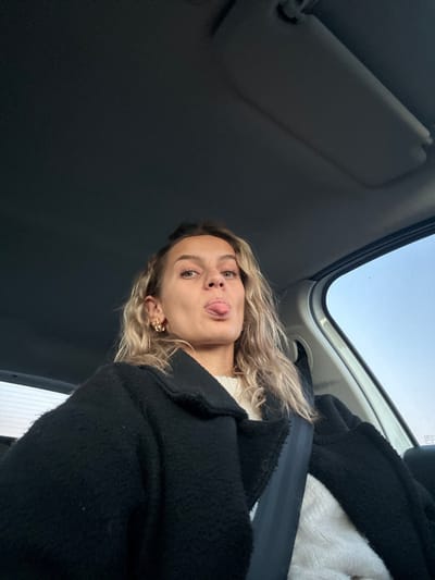 A woman in a car sticks her tongue out towards the camera. She wears a black coat, a white top, and floral earrings. She has fair skin and light blonde, wavy hair. A seatbelt crosses her chest. The vehicle's interior is visible, including a sun visor and a window showing the sky. The image is slightly tilted.